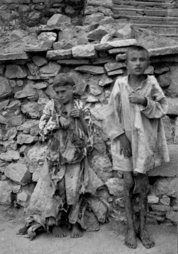 Two children in rags stand at a dry-stone wall. Harpoot, c. 1919-1922.