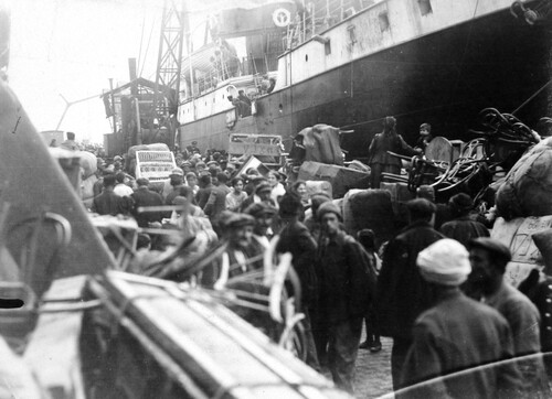 Refugees wait on the quay beneath a steamer's side among bedsteads, chests, and corded bales. Salonica, c. 1922-1923.