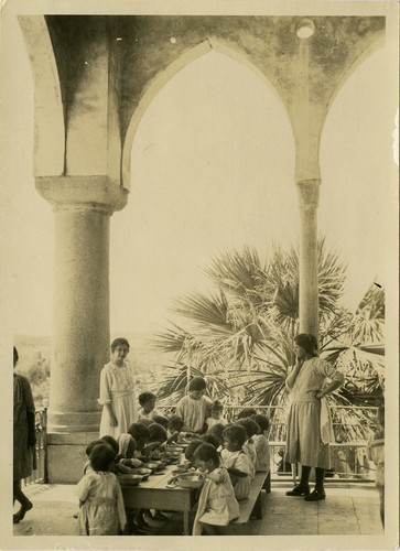 Small children eat from tin bowls at a long table on the Birds' Nest verandah, Sidon, c. 1921–1923.
