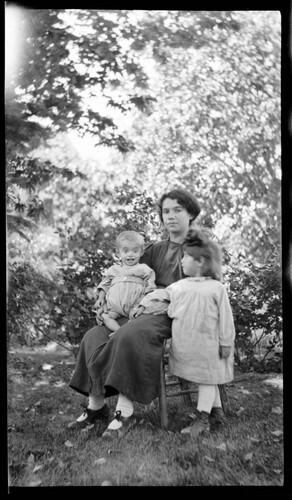 A young woman seated outdoors holds a thin infant while a small girl stands beside her in a leafy garden. Unidentified Near East Relief site.